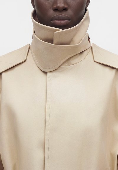 Gros plan d’une personne portant un trench beige avec un col haut structuré et des épaulettes boutonnées.