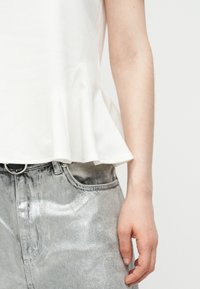 Κοντινό πλάνο ενός ατόμου που φοράει λευκή φαρδιά μπλούζα και ασημένιο μεταλλικό τζιν με μαύρη ζώνη ορατή στη μέση.