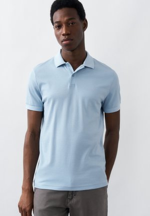 Νέος άνδρας που φοράει ανοιχτό μπλε πόλο πουκάμισο με κοντά μανίκια και γκρι παντελόνια, στέκεται μπροστά σε απλό λευκό φόντο.