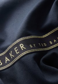 Donkerblauwe stof met een gladde textuur, voorzien van een lint met gouden accenten waarop duidelijk "BAKER by TED BAKER" staat.