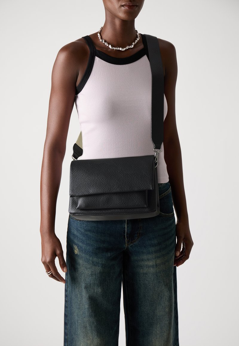 Sac bandoulière en cuir noir avec une surface texturée et un rabat. Bandoulière détachable et design minimaliste. Porté avec un jean en denim.