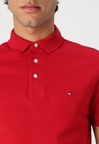 Gros plan d’une personne portant un polo rouge à trois boutons avec un petit logo rectangulaire sur la poitrine.