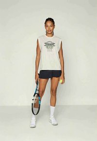 Noor naine, kellel on seljatagune valge top lillemustriga ja must tenniseseelik, hoides tennisereketti ja -palli, seistes tasasel taustal.