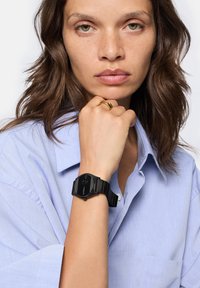 Digitaluhr mit schwarzem Harzarmband und rechteckigem Zifferblatt, ausgestattet mit einem dunklen Display. Getragen an einer Hand mit einem goldenen Ring und einem blauen Hemd.