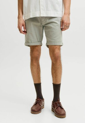 Mand iført lysegrå shorts med opkrogede ben, mørkebrune sokker og brune læder snørestøvler stående mod en ensfarvet baggrund.