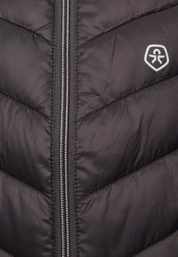 Crna pufasta jakna s chevron uzorkom. Ima prednji zip i mali logo u gornjem lijevom kutu. Glatka tekstura.