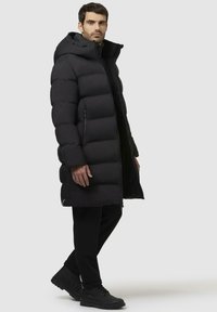 Sort pufferjakke med hætte, der har en quilted design, front lynlåslukning, sidelommer og en længde til knæene. Bæres over sorte bukser og støvler.