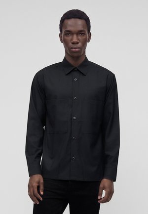 Jeune homme portant une chemise noire à boutons avec deux poches poitrine, se tenant devant un fond gris clair uni.