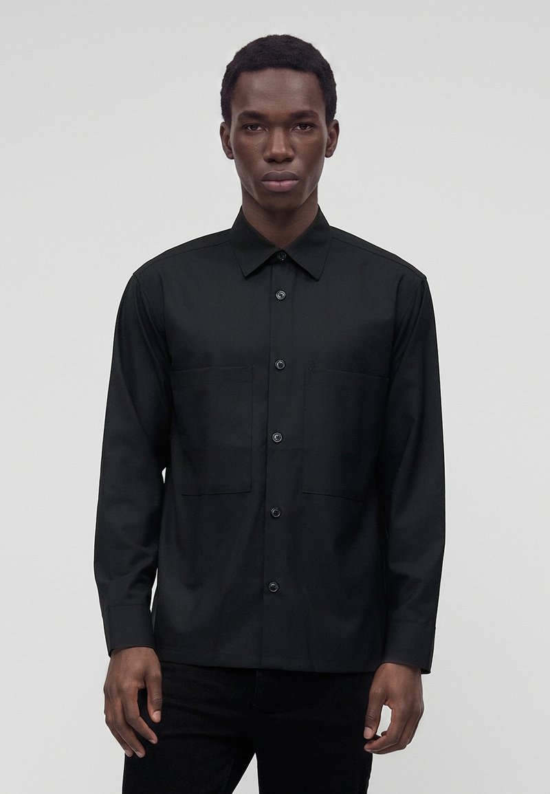 Jeune homme portant une chemise noire à boutons avec deux poches poitrine, se tenant devant un fond gris clair uni.