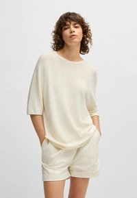 Loose-fitting, cream-colored knit top with short sleeves, smooth texture, and wide neckline, paired with light-colored shorts.