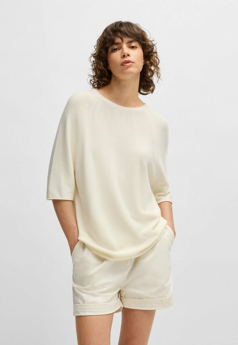 Loose-fitting, cream-colored knit top with short sleeves, smooth texture, and wide neckline, paired with light-colored shorts.