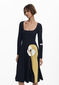 Schwarzes langärmeliges Kleid mit quadratischem Ausschnitt, das auf dem Rock mit einem großen abstrakten Blumenmuster in Gelb, Weiß und Burgunderrot verziert ist.