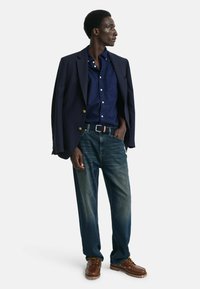 Blazer blu navy con bottoni dorati, camicia blu scura, jeans sbiaditi e scarpe con lacci marroni. L'individuo è in piedi con una mano in tasca.