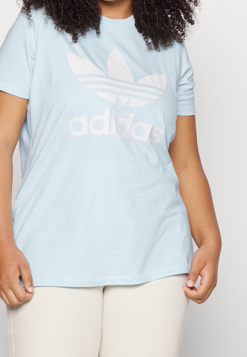 Ljusblå bomulls-t-shirt med en stor vit Adidas-logotyp med tre ränder ovanför ordet "adidas". Kortärmad och avslappnad passform.