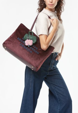 Grande borsa tote in velluto borgogna con testo ricamato, abbellita da un portachiavi verde e rosa. Presenta una maniglia superiore robusta e piedi in ottone.