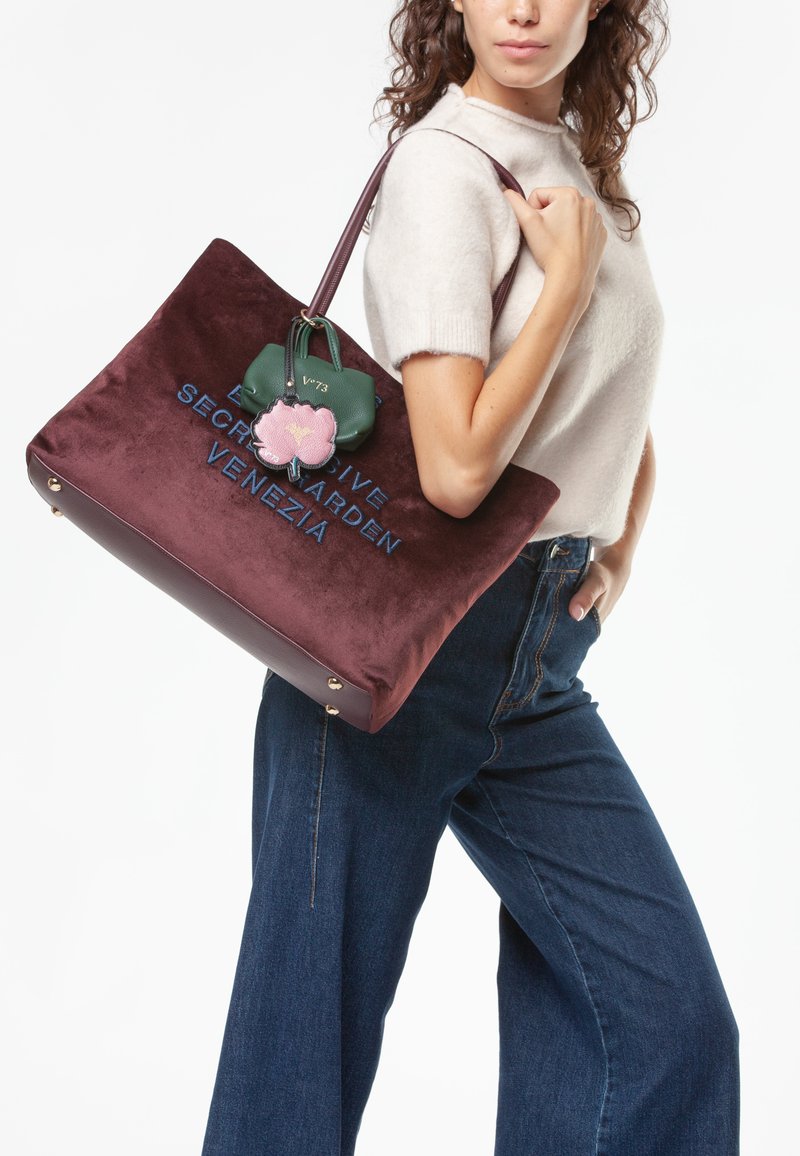 Grande borsa tote in velluto borgogna con testo ricamato, abbellita da un portachiavi verde e rosa. Presenta una maniglia superiore robusta e piedi in ottone.