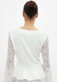 Blouse blanche à manches longues en dentelle avec broderies florales et un ourlet peplum. Tissu lisse avec un motif délicat et texturé.