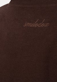 Gros plan sur un tissu marron côtelé avec un texte en lettres cursives brodées discrètement "smilodox" près de l'encolure.