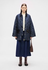 Tengerészkék steppelt kabát fekete gallérral, két elejére varrt zsebbel és arany színű kiegészítőkkel, krémszínű felső és hosszú farmer szoknya fölött barna csizmával viselve.