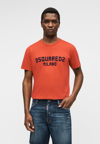 Άνδρας με πορτοκαλί μπλούζα DSQUARED2 Milano και μπλε τζιν σορτς, στέκεται μπροστά σε απλό ανοιχτό γκρι φόντο.