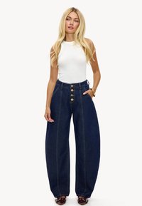 Pantalones anchos de mezclilla oscura con cintura alta, cierre delantero de botones y dos bolsillos laterales, combinados con una camiseta blanca ajustada sin mangas.