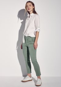 Valkoinen napillinen paita, jonka hihat on käännetty, yhdistetty vaaleanvihreisiin slim-fit-housuihin ja valkoisiin lenkkareihin, joissa on beige yksityiskohtia, taustana yksiön väri.