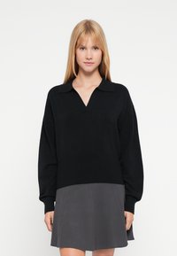 Pulover negru tricotat cu gât în V și guler, cu mâneci lungi și croială lejeră. Materialul pare moale și texturat.