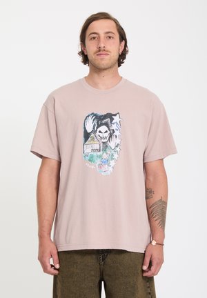Homme portant un t-shirt rose pâle surdimensionné avec un motif représentant un crâne, une maison, une personne qui fait signe de la main et des étoiles, associé à un pantalon marron et un tatouage sur le bras.