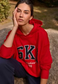 Sudadera oversized roja con "SKI" en grandes letras negras, con capucha forrada, puños acanalados y un corte relajado. Pantalones oscuros visibles.