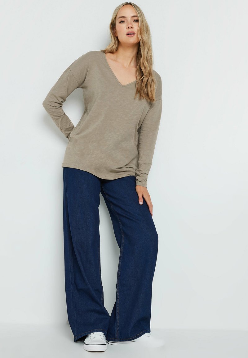 Beige langemouwen V-hals top gecombineerd met blauwe wijde denim broek en witte sneakers, met een relaxte pasvorm en zachte textuur.