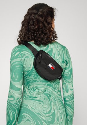 Musta nyloni vyölaukku, jossa on etuvetoketju ja logo, jossa on punainen, valkoinen ja sininen väri; laukku on puettu vartalon yli vihreän kuvioidun yläosan päällä.