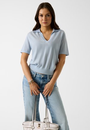 Frau trägt hellblaues Kurzarmoberteil und Jeans, hält eine beige Handtasche an den Griffen vor weißem Hintergrund.