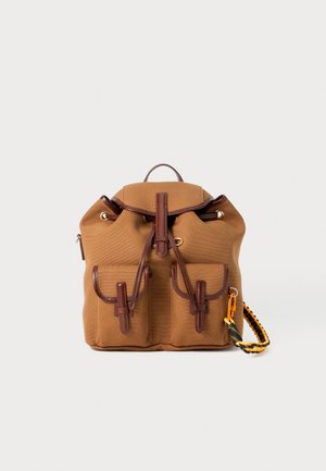 Sac à dos en tissu beige avec des accents en cuir marron, deux poches frontales, fermeture à cordon, et une poignée supérieure. Présente un matériau texturé et une sangle détachable.
