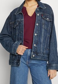 Giacca in denim blu scuro con taglio classico, dotata di apertura con bottoni, due tasche sul petto e una texture leggermente vissuta.