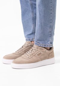 Sneakers in suede beige con punta arrotondata, lacci piatti e suola in gomma bianca. Cuciture evidenti e un piccolo logo decorativo sul lato.