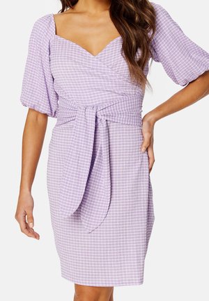 Woman wearing a lilac plaid wrap dress with puff sleeves and a tied waist belt, posing with one hand on her hip.