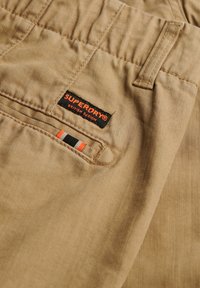 Beige katoenen stoffen broek met een gestikte textuur, voorzien van een zwart label met "SUPERDRY" en rode, zwarte en witte accentdetails.