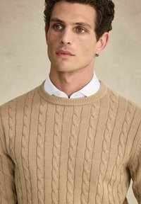 Beige kabelgebreide trui met een ronde halslijn, gemaakt van een zacht materiaal, met gestructureerde, verhoogde patronen langs het lichaam.