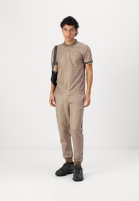 Beige Poloshirt und Jogginghose mit einem Rautenmuster, schwarzem Saum und seitlichen Streifen, kombiniert mit schwarzen Sportschuhen. Schultertasche inklusive.