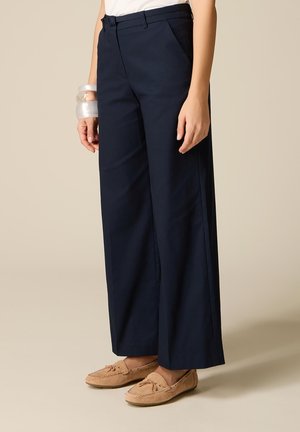 Donna che indossa pantaloni larghi blu navy, mocassini marrone chiaro e grandi braccialetti trasparenti al polso sinistro, su sfondo beige.