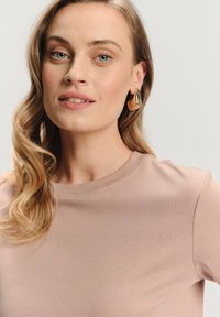 Blondynka z luźnymi lokami, ubrana w beżową bluzkę i duże, złote geometryczne kolczyki, lekko patrzy w bok na jednolitym tle.