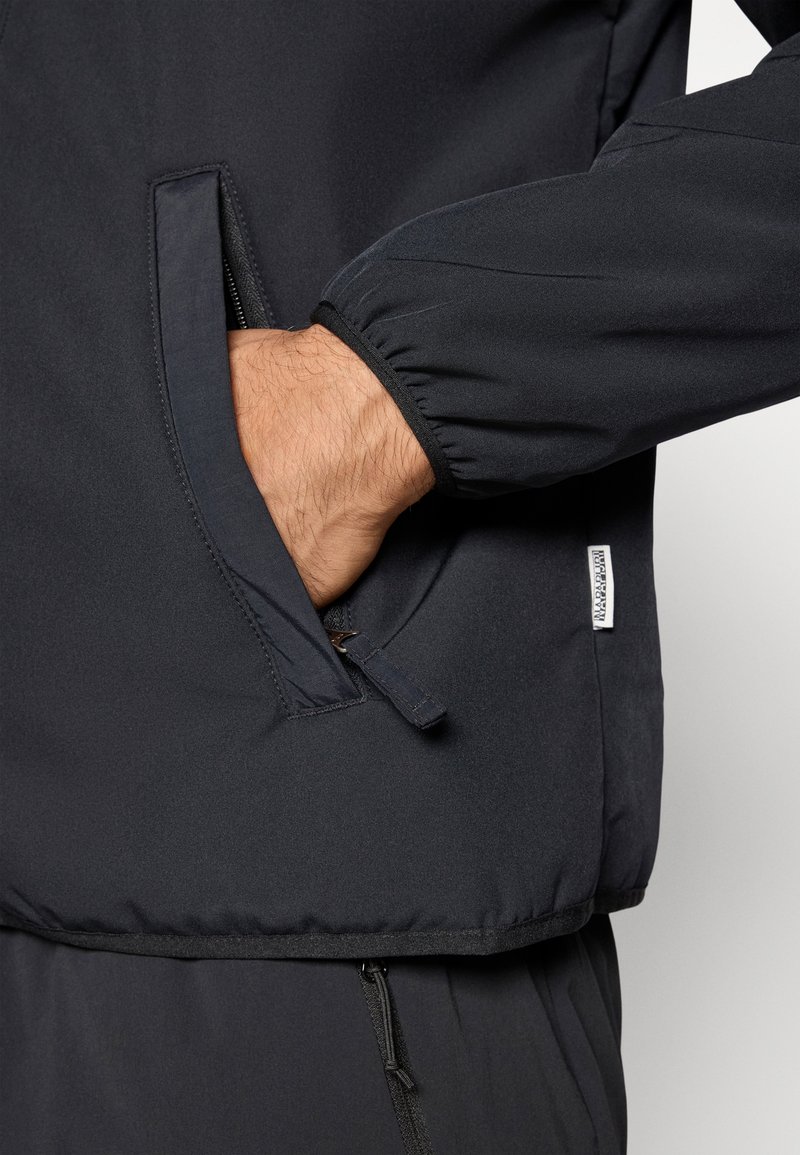 Veste noire avec une texture lisse, dotée d'une poche zippée et de poignets élastiques. Étiquette visible sur le côté gauche de la veste.