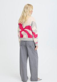 Frau mit langen blonden Haaren, die einen abstrakt gemusterten Pullover in Pink, Grau und Weiß sowie lockere graue Jeans trägt und seitlich steht.