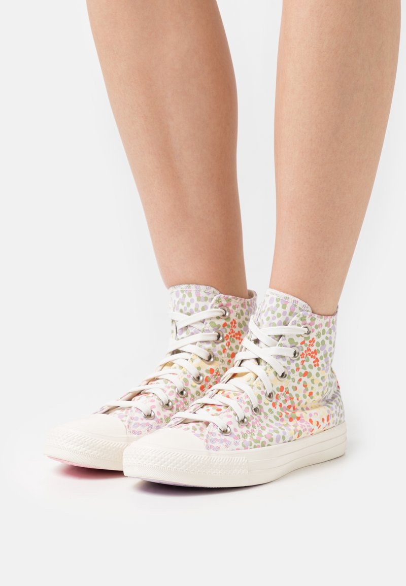 Personne portant des baskets montantes blanches avec un motif floral coloré et à pois sur un fond clair.