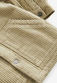 Tissu en velours côtelé beige avec des stries verticales, comportant une poche cousue et deux boutons métalliques ronds avec du texte gravé.