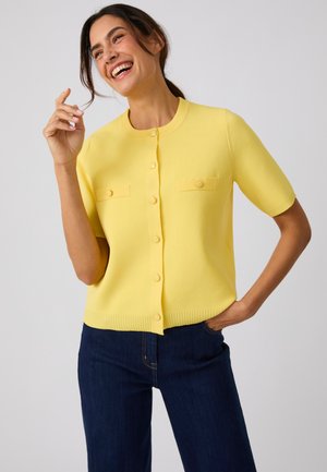 Femme souriante, debout avec une main dans la poche, portant un cardigan jaune à manches courtes boutonné et un jean bleu foncé sur fond uni.