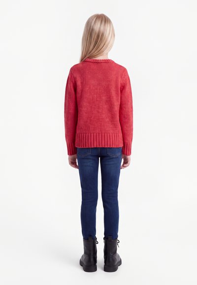 Enfant aux longs cheveux blonds portant un pull rouge, un jean bleu et des bottes noires, debout de dos sur un fond blanc.