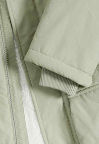 Giacca verde chiaro con un esterno liscio, polsini a costine e chiusura con zip. Foderata con un materiale morbido e testurizzato per il calore.