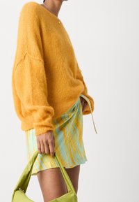 Suéter de mohair amarillo oversize combinado con una falda de colores azul claro y amarillo con estampado. Sosteniendo un bolso de satén verde en la mano derecha.