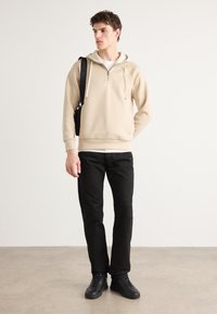 Sweatshirt à capuche beige avec fermeture éclair, doté d'une poche kangourou et de cordons de serrage, associé à un pantalon droit noir et des baskets noires.